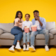 joyful-black-family-three-sitting-sofa-watching-movie-eating-popcorn-from-bucket-isolated-yellow-wall (1)